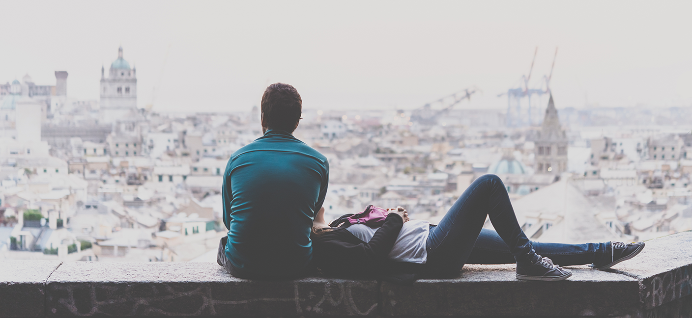 A man sitting with his back to us, watching the city skyline. A woman rests her head on his lap.